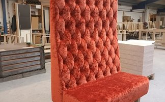 Eine hohe, gepolsterte Bank in leuchtendem Orange steht in einem Werkstattraum voll von Möbelstücken und Holzplatten.