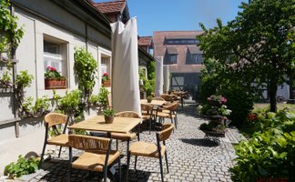 Die Terrasse eines Restaurants ist mit rustikalen Tischen und Stühlen gestaltet, umgeben von blühenden Pflanzen und historischer Architektur.
