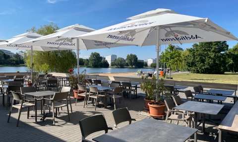 Die Terrasse des Cafés bietet Blick auf den See, umgeben von grünen Bäumen und modernen Gebäuden im Hintergrund.