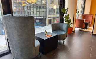Helle Sessel, ein dunkler Tisch und eine grüne Pflanze gestalten die einladende Ecke des Cafés bei Tageslicht.