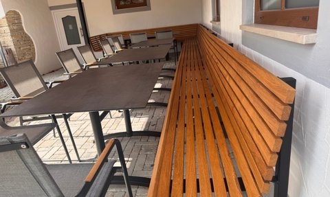 In einem gemütlichen Gartenbereich sind eine Holzbank und mehrere Stühle um einen großen Tisch angeordnet, bereit für Gäste.