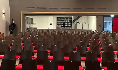 Reihen von leeren Stühlen in einem Kino, die auf eine Bühne zeigen, umgeben von einem modernen, stilvollen Raum mit rotem Akzent.