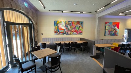 Ein gemütlicher Restaurantbereich mit Holzstühlen und Tischen, umgeben von lebhaften Kunstwerken und einem großen Eingang.