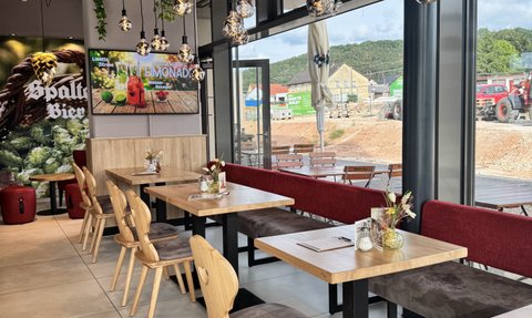 Die Inneneinrichtung des Cafés zeigt helle Holzmöbel, gemütliche Sitzgelegenheiten und eine große Fensterfront mit Ausblick.