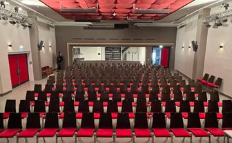 Reihen von leeren Stühlen in einem Kino, die auf eine Bühne zeigen, umgeben von einem modernen, stilvollen Raum mit rotem Akzent.