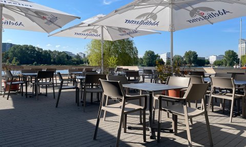 Modern gestalteter Cafébereich am Wasser, abgedeckt mit großen Sonnenschirmen und zahlreichen Tischen und Stühlen.