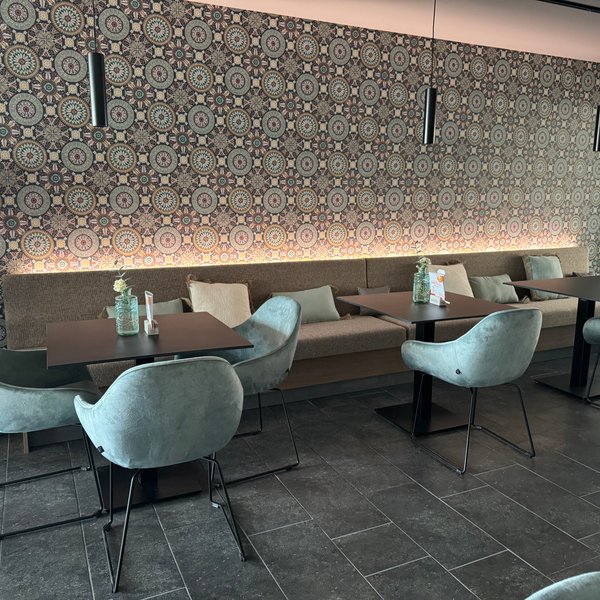 In einem modernen Café stehen runde Tische mit gemütlichen, geschwungenen Stühlen, umgeben von einem dekorativen Wandmuster.