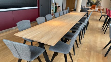 Ein langer Holzttisch steht in einem modernen Raum, umgeben von mehreren grauen Stühlen mit schwarzen Beinen.