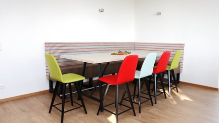 Sitzecke mit bunter Bank und fünf Stühlen in grün, rot und hellblau sowie einem weißen Tisch