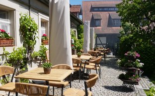 Die Terrasse eines Restaurants ist mit rustikalen Tischen und Stühlen gestaltet, umgeben von blühenden Pflanzen und historischer Architektur.