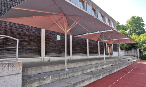 Drei große Sonnenschirme stehen auf einer Treppe einer Schule, mit Holzfassade im Hintergrund und rotem Sportplatz.