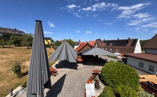 Der sonnige Garten eines Gasthauses mit großen Sonnenschirmen, Holzstühlen und einer ruhigen Atmosphäre unter blauem Himmel.