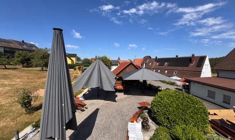 Der sonnige Garten eines Gasthauses mit großen Sonnenschirmen, Holzstühlen und einer ruhigen Atmosphäre unter blauem Himmel.