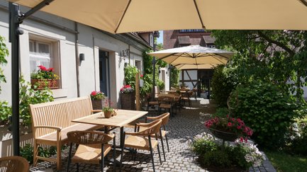 Die Terrasse eines Restaurants ist mit rustikalen Tischen und Stühlen gestaltet, umgeben von blühenden Pflanzen und historischer Architektur.