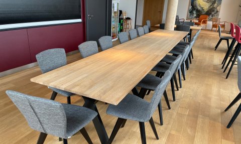 Ein langer Holzttisch steht in einem modernen Raum, umgeben von mehreren grauen Stühlen mit schwarzen Beinen.