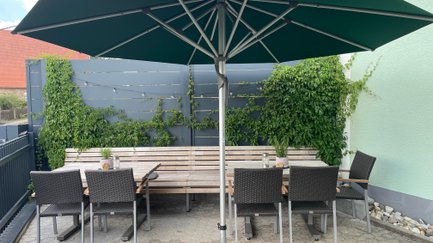 Ein stilvoller Außenbereich mit einem langen Tisch, bequemen Stühlen und grünem Sonnenschirmn, der zum Verweilen einlädt.