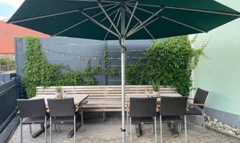 Ein stilvoller Außenbereich mit einem langen Tisch, bequemen Stühlen und grünem Sonnenschirmn, der zum Verweilen einlädt.
