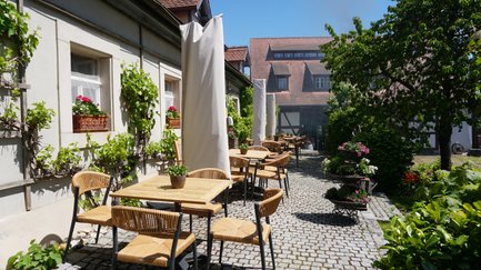 Die Terrasse eines Restaurants ist mit rustikalen Tischen und Stühlen gestaltet, umgeben von blühenden Pflanzen und historischer Architektur.