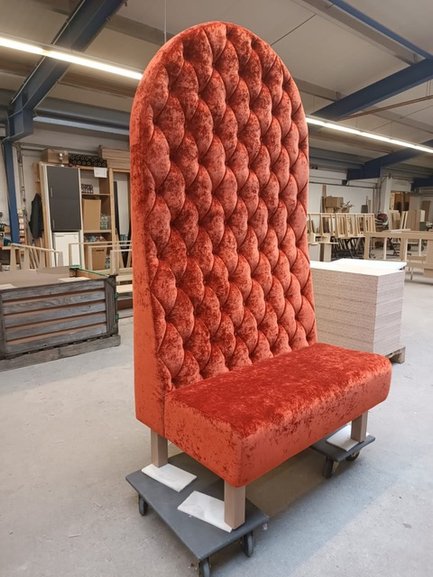 Eine hohe, gepolsterte Bank in leuchtendem Orange steht in einem Werkstattraum voll von Möbelstücken und Holzplatten.