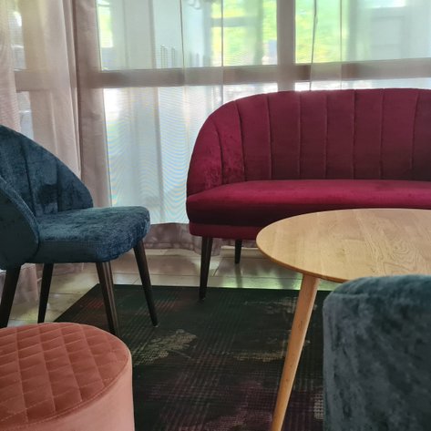 In einem hellen Raum stehen ein rotes Sofa und ein blauer Stuhl, ergänzt durch einen runden Tisch und einen gepolsterten Hocker.