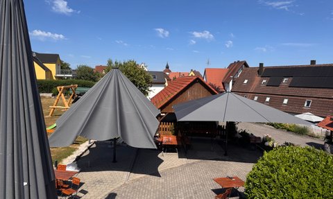 Der Außenbereich eines Gasthofs, ausgestattet mit modernen Sonnenschirmen und rustikalen Holzmöbeln, umgeben von ländlicher Idylle.