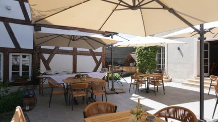 Die Terrasse eines Restaurants ist mit rustikalen Tischen und Stühlen gestaltet, umgeben von blühenden Pflanzen und historischer Architektur.