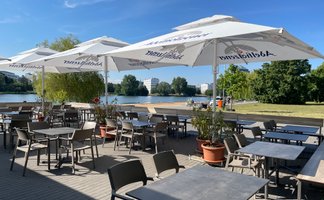 Die Terrasse des Cafés bietet Blick auf den See, umgeben von grünen Bäumen und modernen Gebäuden im Hintergrund.