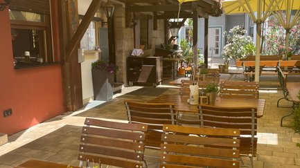 Sonnige Terrasse eines Restaurants, bestückt mit Tischen und Stühlen, bereit für Gäste, die das schöne Wetter genießen wollen.