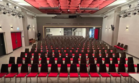 Reihen von leeren Stühlen in einem Kino, die auf eine Bühne zeigen, umgeben von einem modernen, stilvollen Raum mit rotem Akzent.
