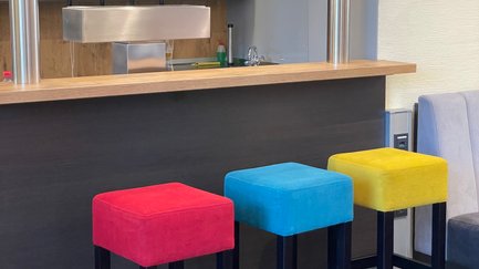Drei farbenfrohe Barhocker in Rot, Blau und Gelb stehen an einer modernen Bar mit Gläsern im Hintergrund.