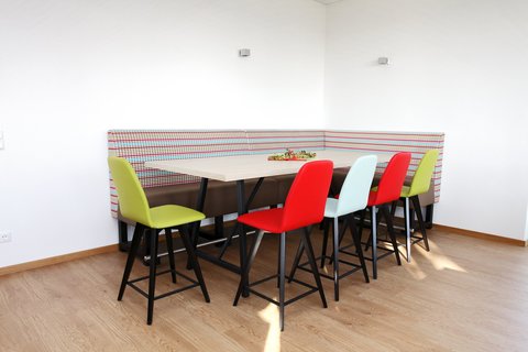 Sitzecke mit bunter Bank und fünf Stühlen in grün, rot und hellblau sowie einem weißen Tisch